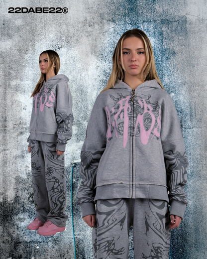 A model wearing the Grey/Pink Jogger from 22DABE22, a streetwear brand. The jogger is made from a blend of cotton and polyester, with a peached interior for comfort. The jogger features screen print designs on both sides and an embroidery appliqué at the back pockets.