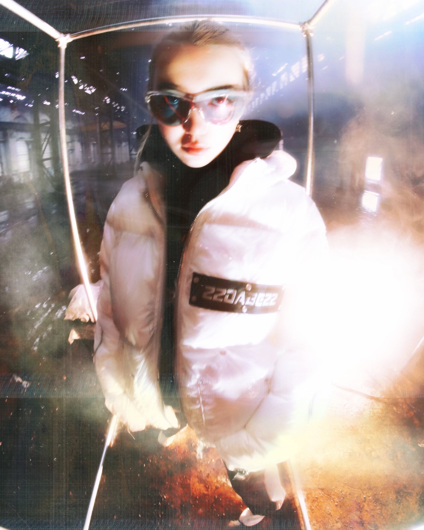 A white puffer jacket from 22DABE22, made of high-quality nylon fabric and cotton down filling, featuring custom logo rivets and buttons, detachable hood, and adjustable drawcords on the bottom hem. The jacket has an oversized fit, with sleeves that can be unzipped for unrestricted movement and ample storage space with one inside pocket and two side pockets. The logo tape at the chest adds a touch of branding.