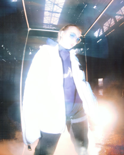 A white puffer jacket from 22DABE22, made of high-quality nylon fabric and cotton down filling, featuring custom logo rivets and buttons, detachable hood, and adjustable drawcords on the bottom hem. The jacket has an oversized fit, with sleeves that can be unzipped for unrestricted movement and ample storage space with one inside pocket and two side pockets. The logo tape at the chest adds a touch of branding.